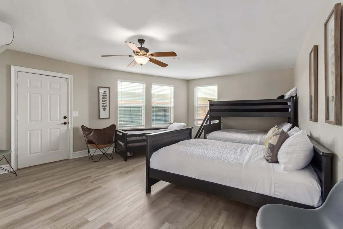 A cheerful, family-friendly bedroom with a bunk bed and two twin beds, large windows letting in morning light, ceiling fan for comfort, and playful artwork on the wall. The room feels welcoming and ideal for children or groups of friends.