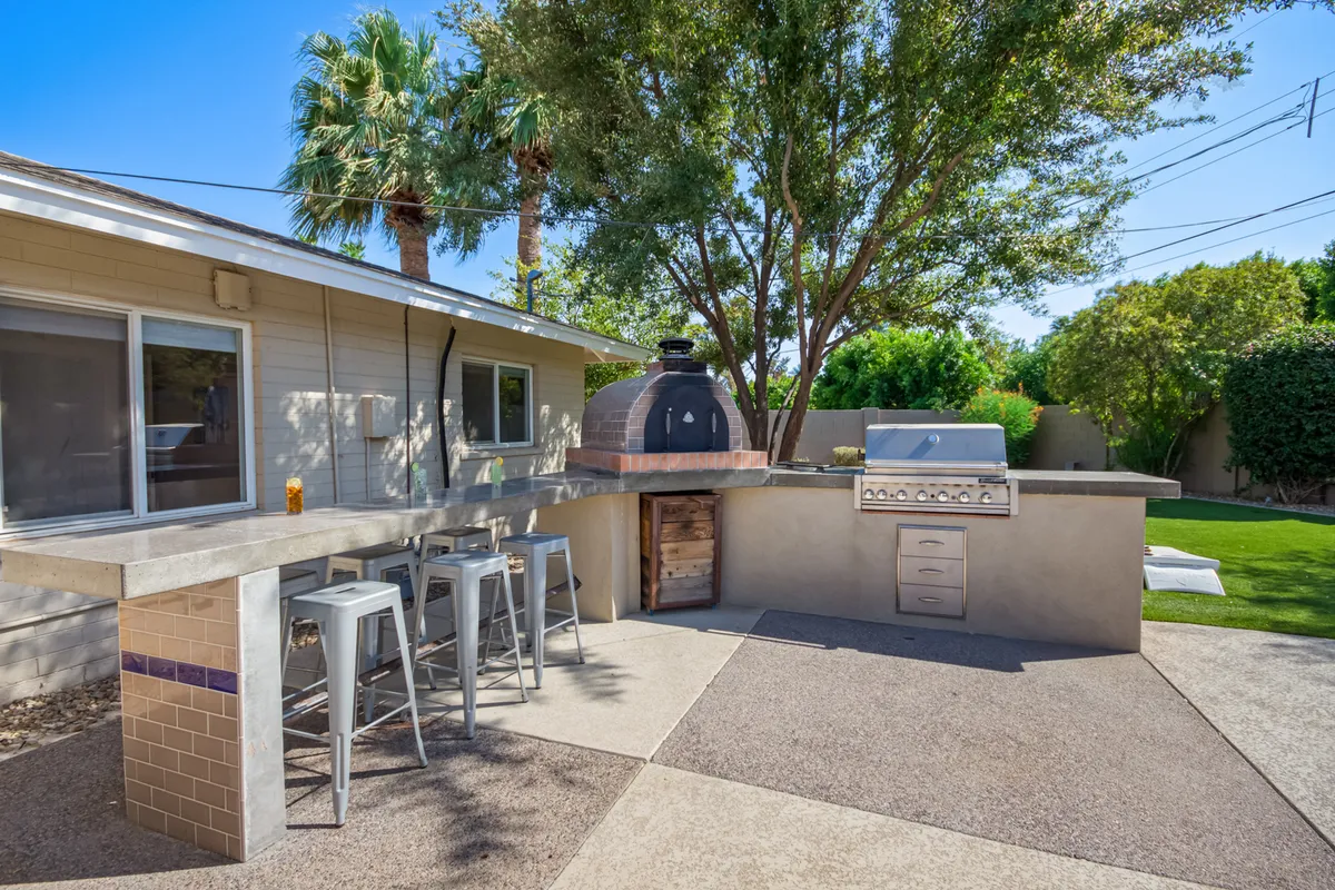 A stylish outdoor kitchen area with a prominent wood-fired pizza oven, grill, bar stools, and palm-framed views. The design invites guests to cook, eat, and socialize in a resort-like setting.