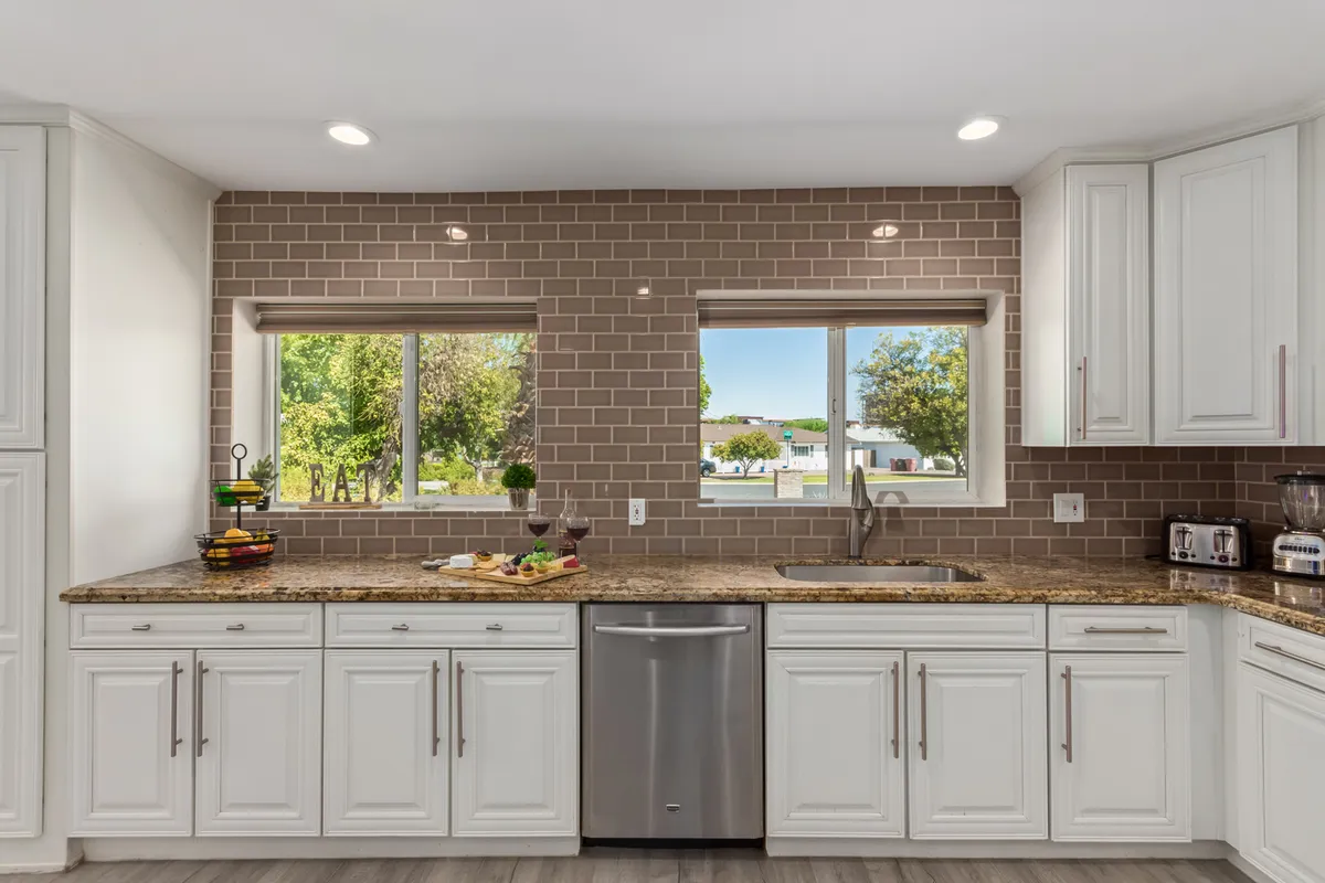 This kitchen image focuses on a granite countertop set before double windows, with modern appliances and a serving tray filled with food. The scene evokes a spirit of hospitality, with light filling the space and plenty of room for meal prep or casual dining.