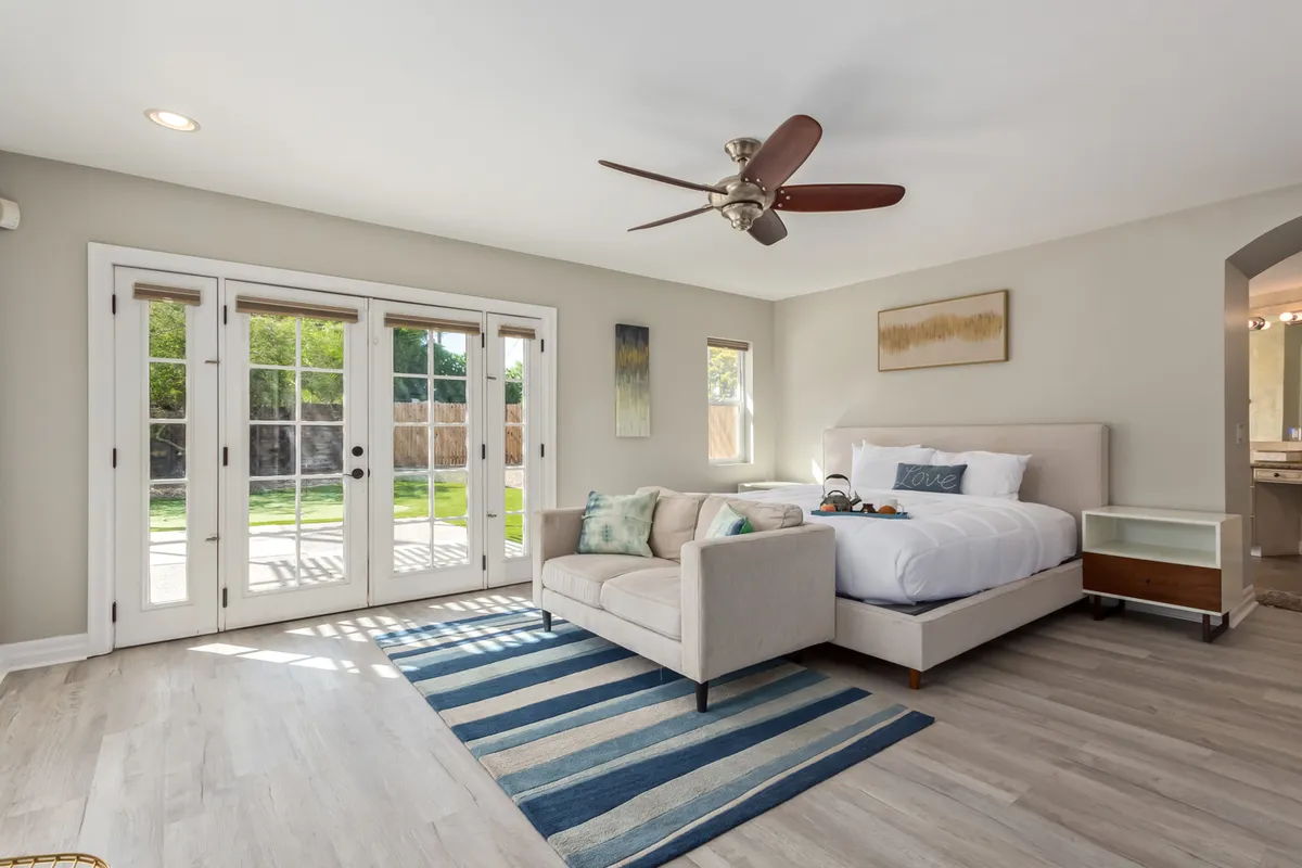 A bright, spacious bedroom featuring a large bed adorned with decorative pillows, a comfortable sofa set against large windows, and a striped rug adding a touch of style. Soft afternoon sunlight filters through the expansive windows, creating a calm and inviting atmosphere.