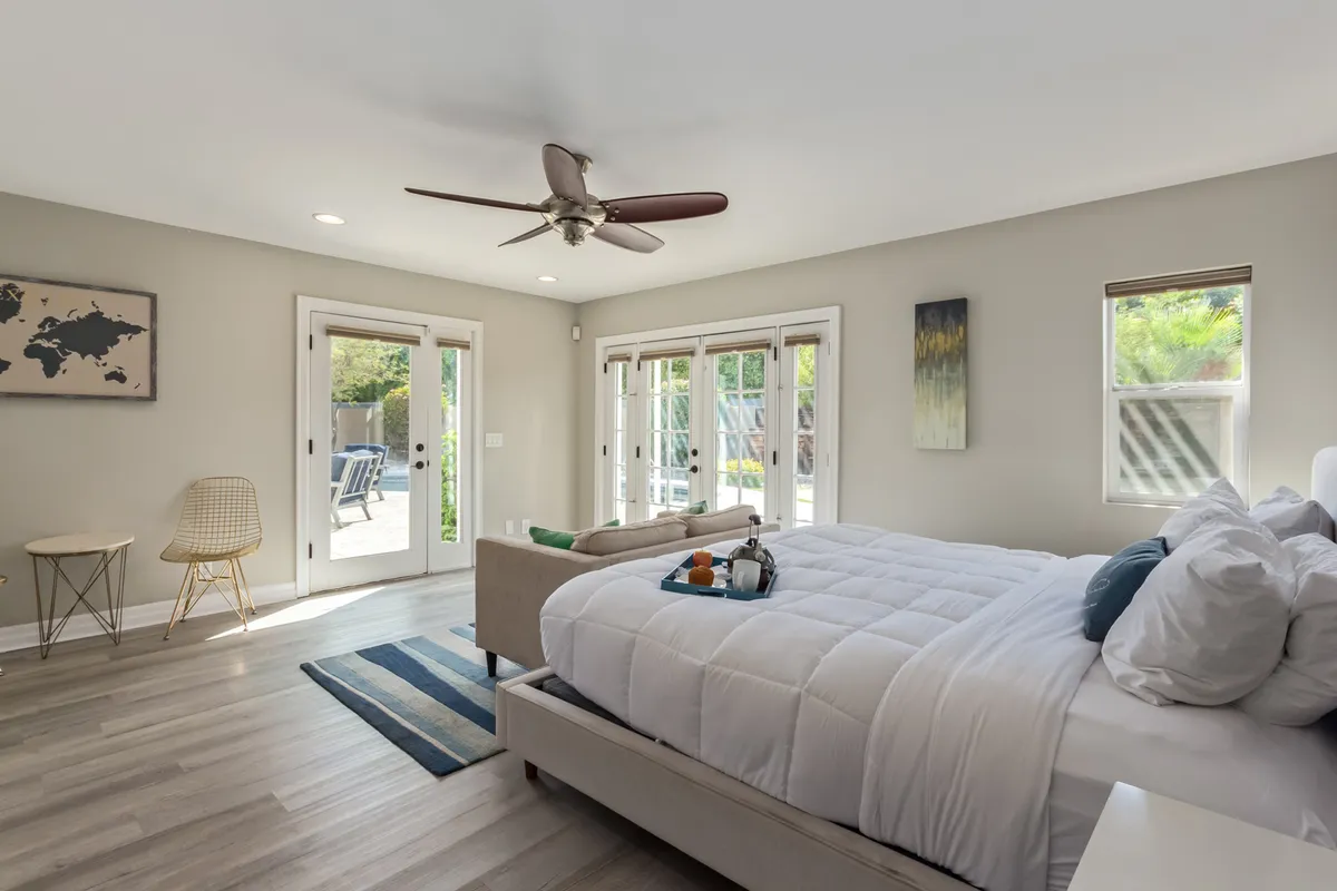 A tranquil bedroom featuring a plush bed with white bedding, two sets of French doors that open onto a scenic view, a comfortable chair, and world map artwork. The abundance of natural light and minimalist decor create a peaceful retreat.