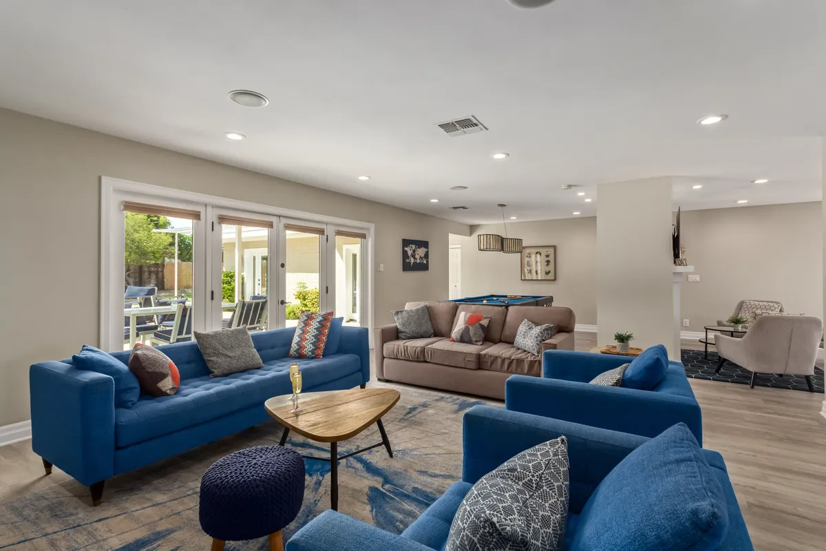 A spacious living room featuring blue sofas arranged around a wooden coffee table, accented with decorative pillows. Large windows line the back wall, flooding the room with natural light and providing glimpses of the surrounding desert landscape. The scene invites gathering and relaxation, ideal for both entertaining and unwinding.