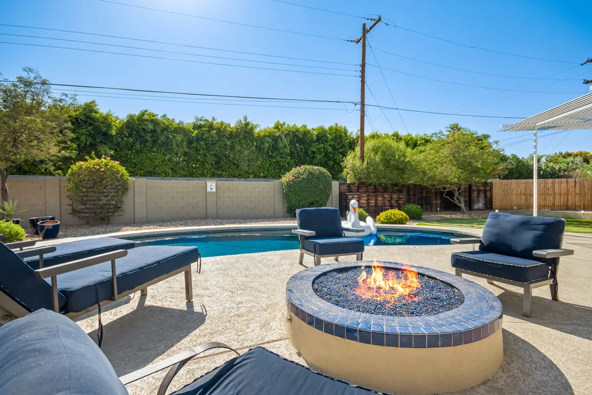 A sunlit backyard scene featuring a sparkling pool, inviting lounge chairs, lush landscaping, and a cozy fire pit beside the water. The setting is perfect for group fun—kids splashing, adults relaxing, and evenings spent toasting marshmallows under the open sky.
