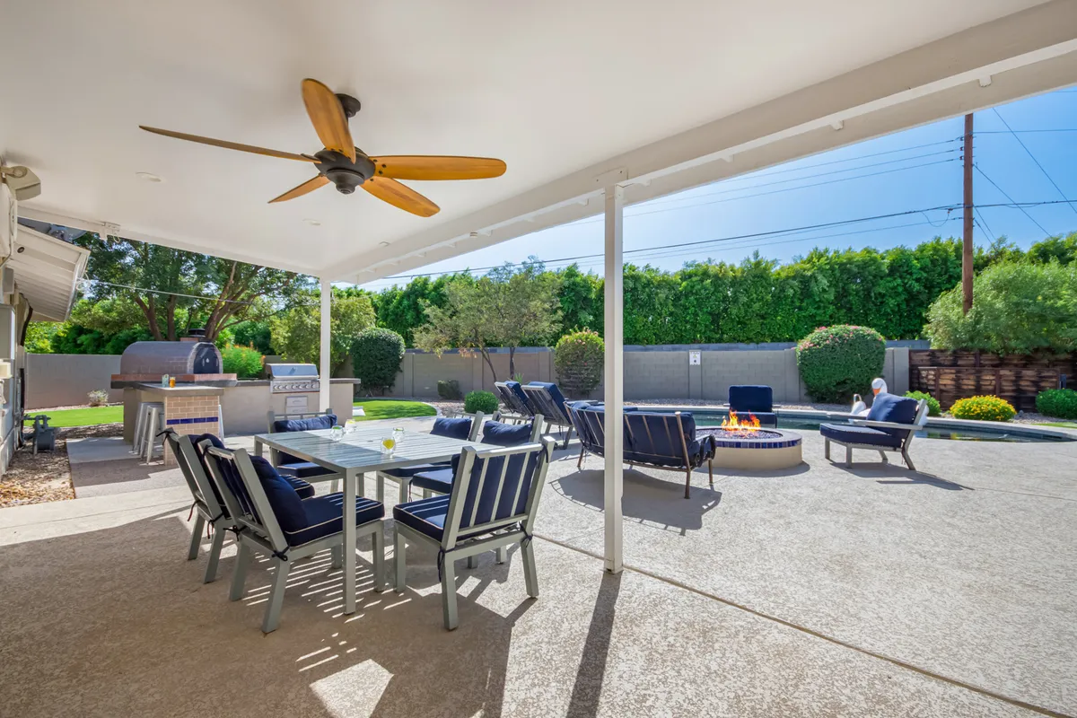 An inviting patio scene featuring a large outdoor dining table, comfortable lounge chairs, a fire pit at center, and an outdoor kitchen area. The setting is perfect for group meals, late-night conversations, and relaxing by firelight.