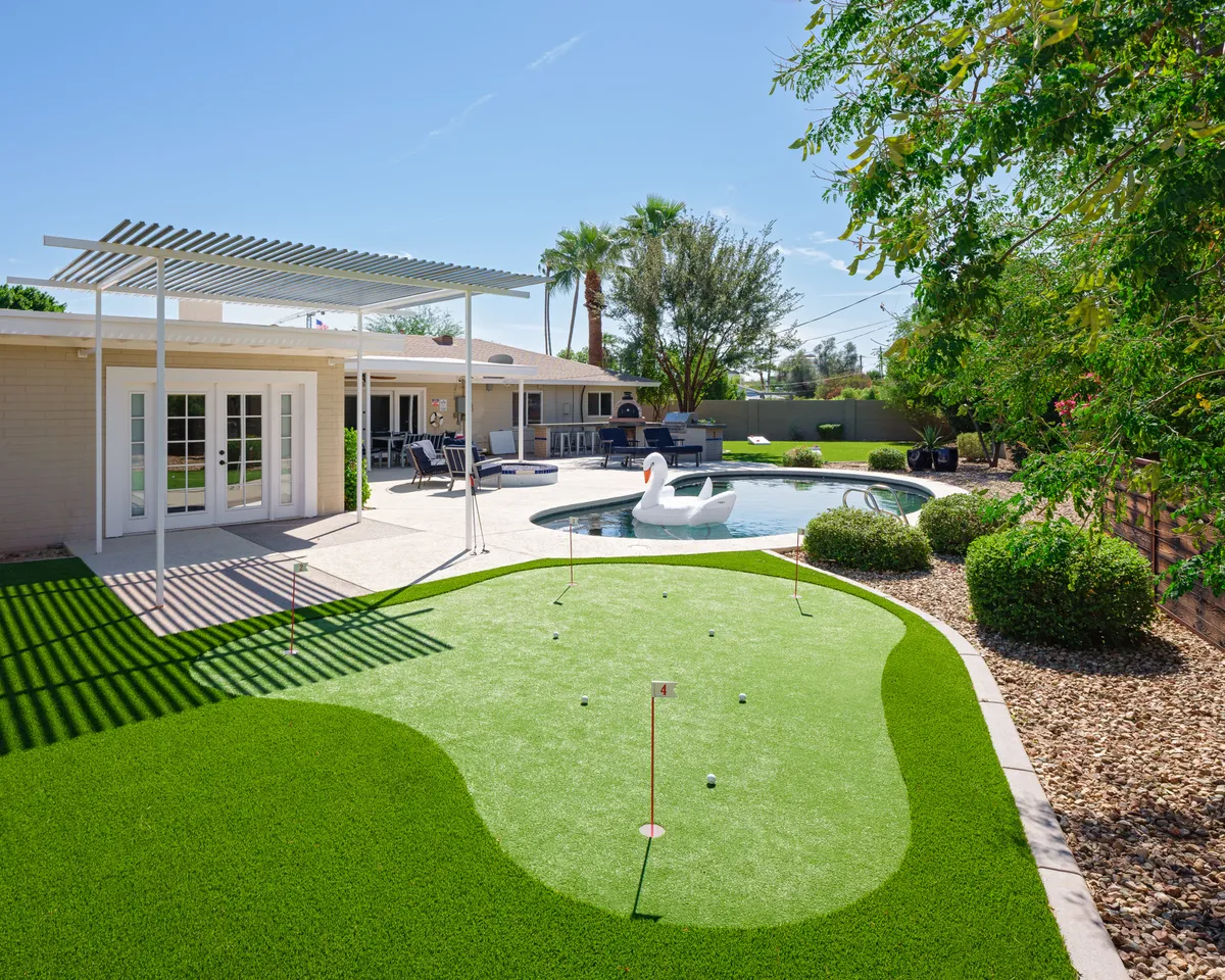 A vibrant exterior view showcasing a sparkling swimming pool, adjacent putting green, a pergola, and a whimsical swan lawn ornament. The area is set up for lounging, sunbathing, golfing, and outdoor dining, all with a backdrop that suggests mountain views and a fun, relaxed atmosphere.