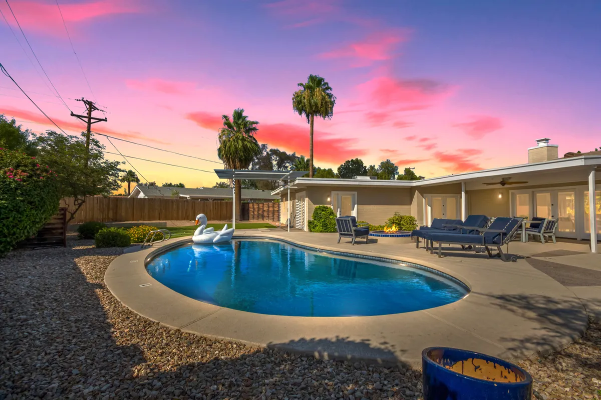 The image showcases a sparkling swimming pool surrounded by sleek lounge chairs and tall palm trees, set against a vibrant, colorful sunset sky. The setting exudes an inviting, relaxed mood, perfect for unwinding, socializing, or enjoying a quiet desert evening. The pool area is the centerpiece, suggesting possibilities for sunset swims, sunbathing, or gatherings under the open Arizona sky, with Camelback Mountain likely visible in the distance.