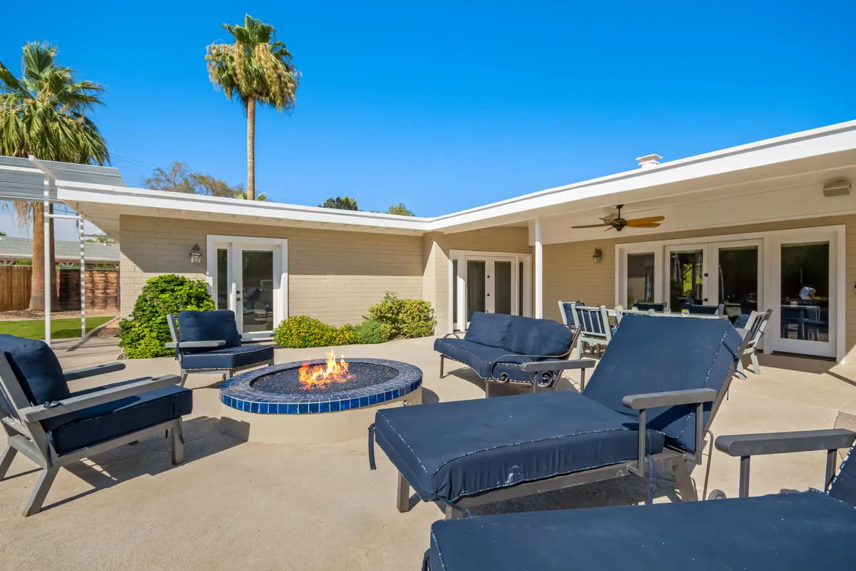 A cozy exterior lounge area featuring a fire pit surrounded by lounge chairs and palm trees, with sliding doors leading back inside. The space is designed for relaxing evenings under the stars, perfect for gathering and enjoying culinary activities at home.